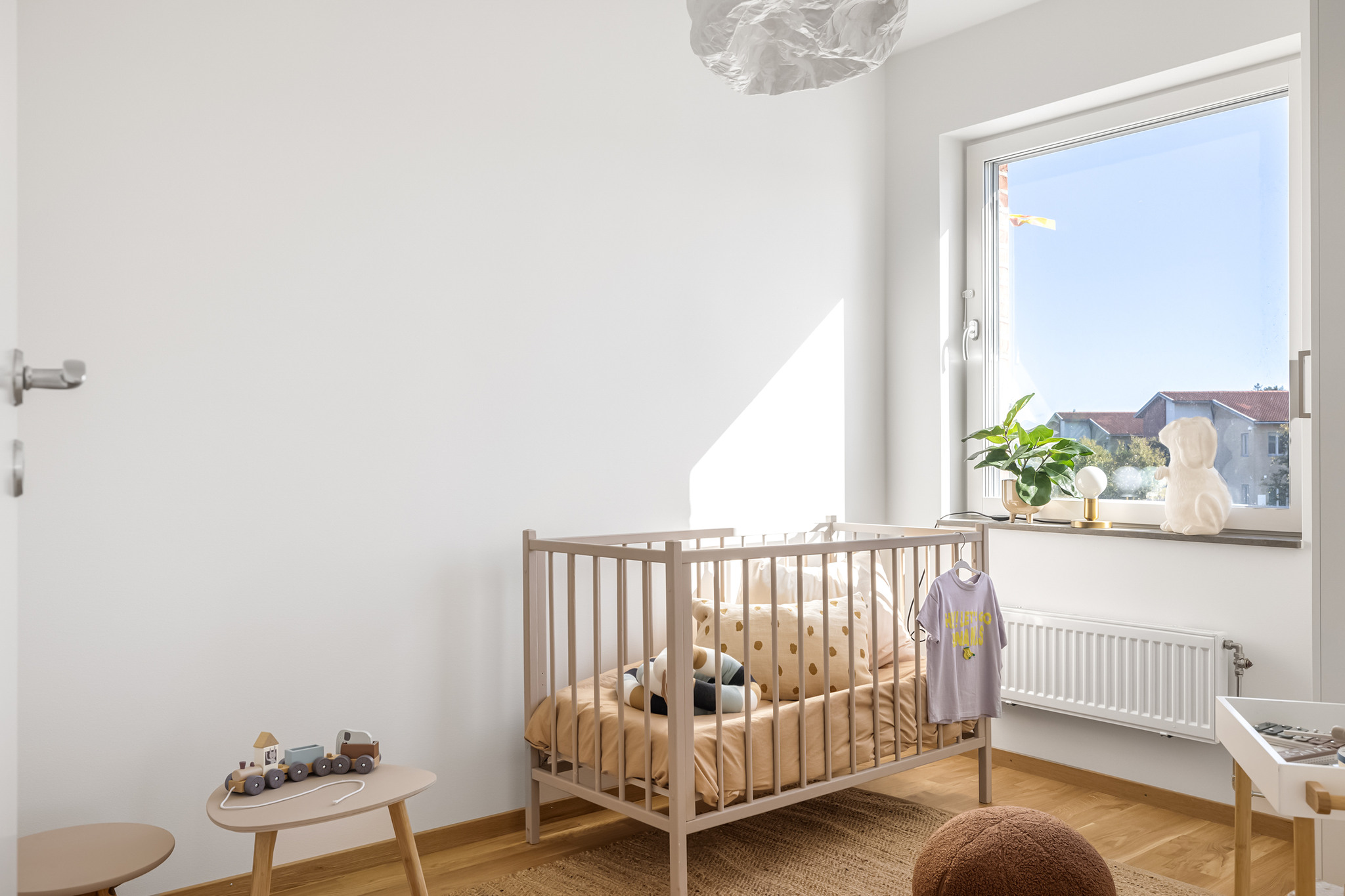 Ett ljust barnrum med spjälsäng och plats för förvaring i garderob. Ett stort fönster ger gott om ljusinsläpp.