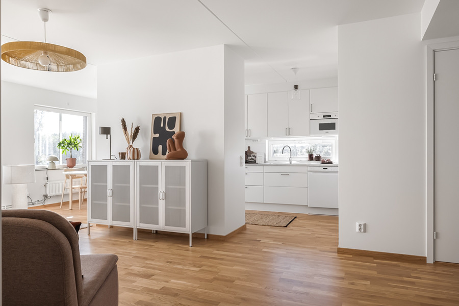 Ljust vardagsrum i vitt som möter ett vitt kök i en öppen planlösning. I anslutning till köks delen finns en matplats och vardagsrums finns plats tv, tv-bänk och sitt ytor.